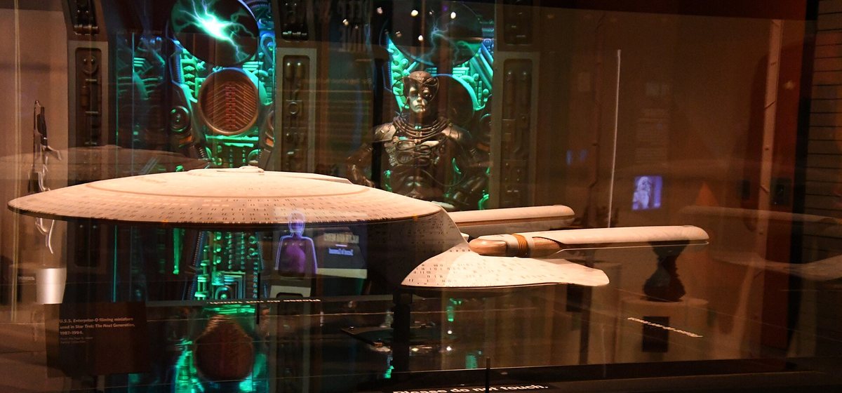Das originale Drehmodell der U.S.S. Enterprise NCC-1701-D aus The Next Generation in einer Museumsvitrine, dahinter ein Borg-Kostüm